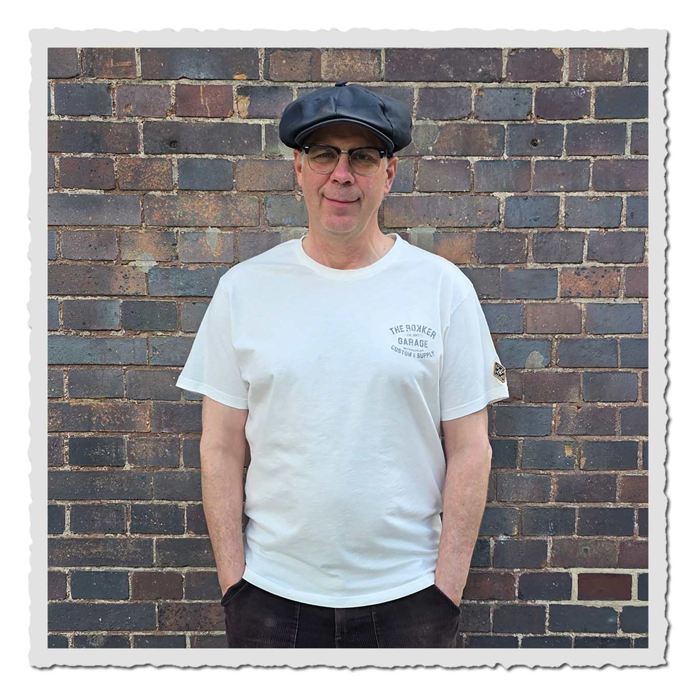 Mann von vorn in hellem T-Shirt mit kleinem Brustprint, dunkler Kappe, vor mehrfarbiger Backsteinwand.